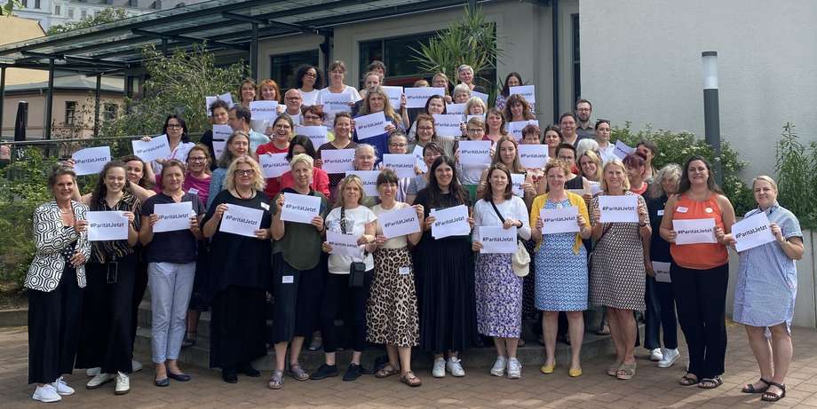 Alle Teilnehmenden der Konferenz halten ein schild mit der Aufschrift "#ParitätJetzt" hoch.