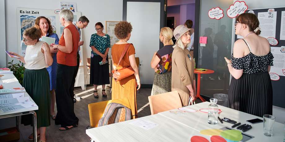 Ein Raum, in dem der Bereich Gleichstellungsberichte ihre Arbeit vorstellt. Es sind einige Mitarbeiterinnen und Besucher*innen im Gespräch.